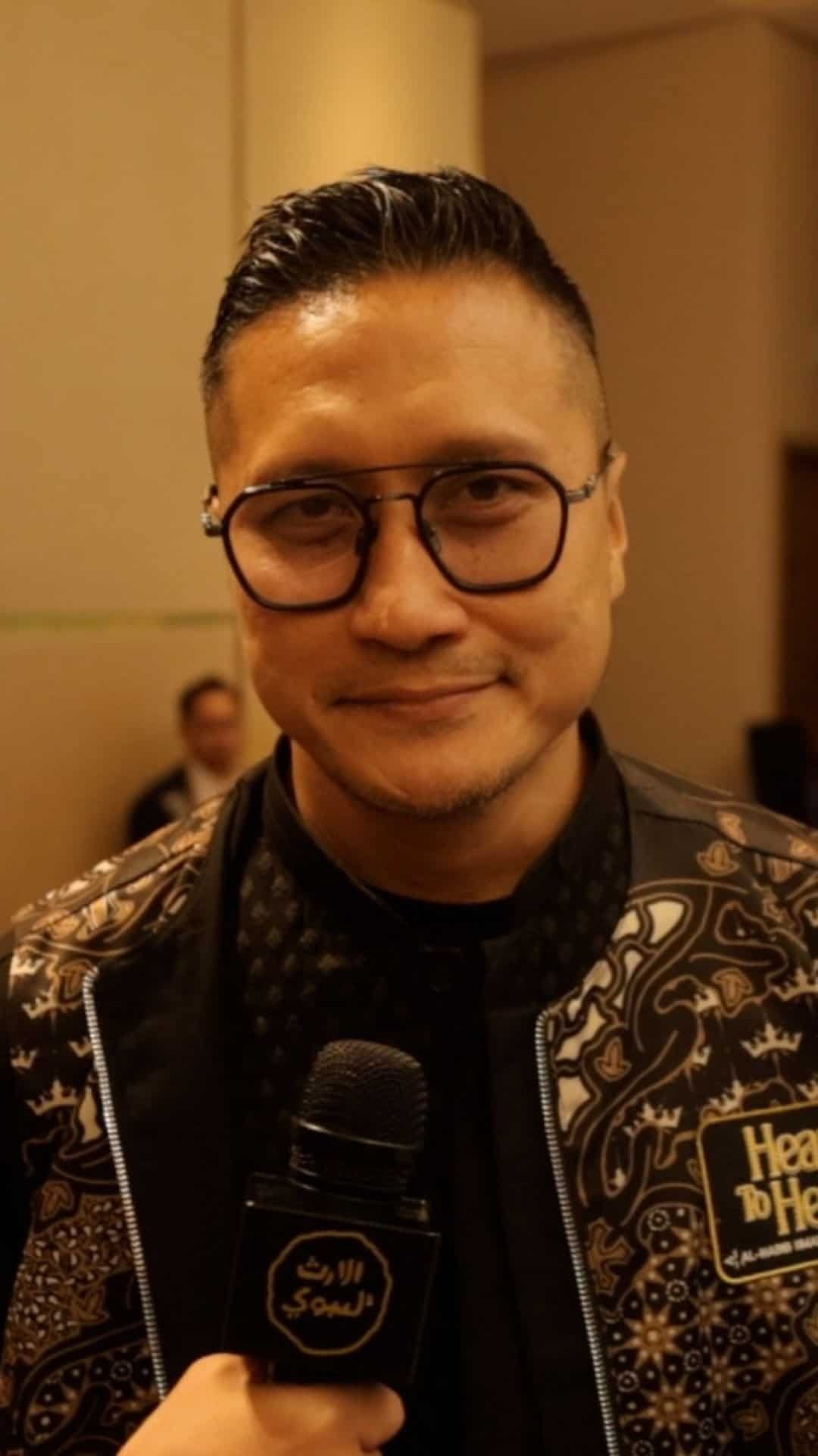 Pencitraan pria profesional dengan kaca mata dan jaket bermotif, berbicara di acara resmi.